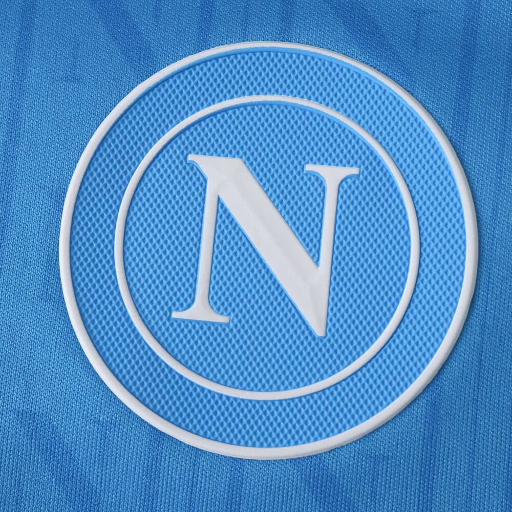 Close-up of the Napoli club crest on the 2025/26 home fan version jersey, featuring a blue background.
