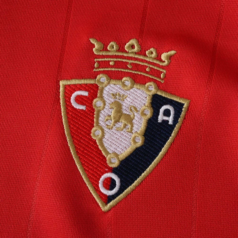 Close-up of Osasuna club crest embroidered on the red 2025/26 home fan jersey fabric.