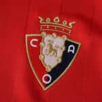Close-up of Osasuna club crest embroidered on the red 2025/26 home fan jersey fabric.