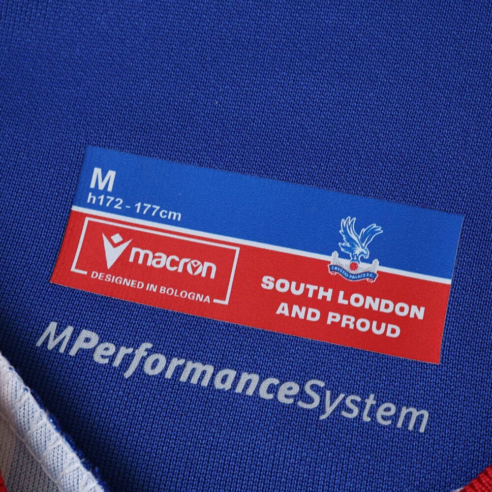 Close-up of Crystal Palace 2025/26 home fan jersey tag with Macron logo and "South London and Proud" text.
