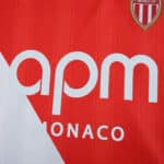 Close-up of the AS Monaco 2025/26 home fan jersey showing club crest and sponsor logo on red and white fabric.