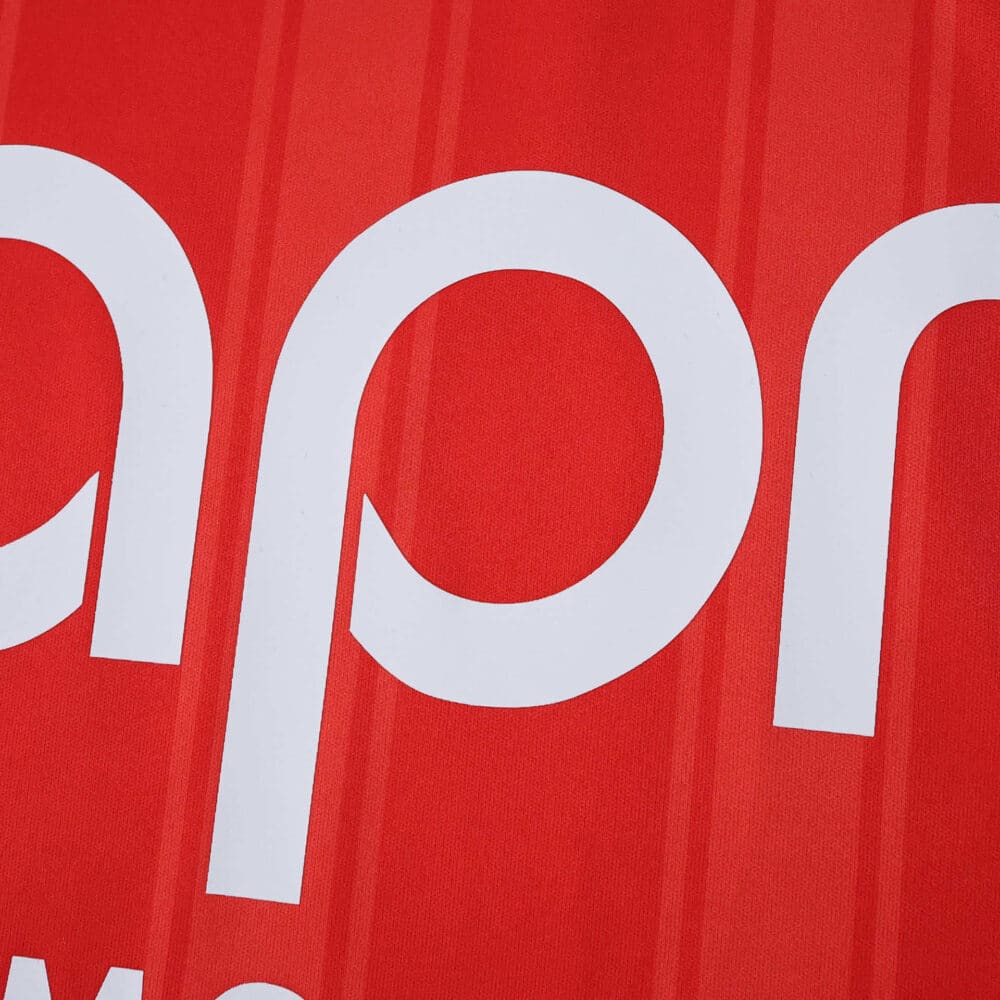 Close-up of AS Monaco 2025/26 home jersey, showing red fabric with white sponsor lettering.